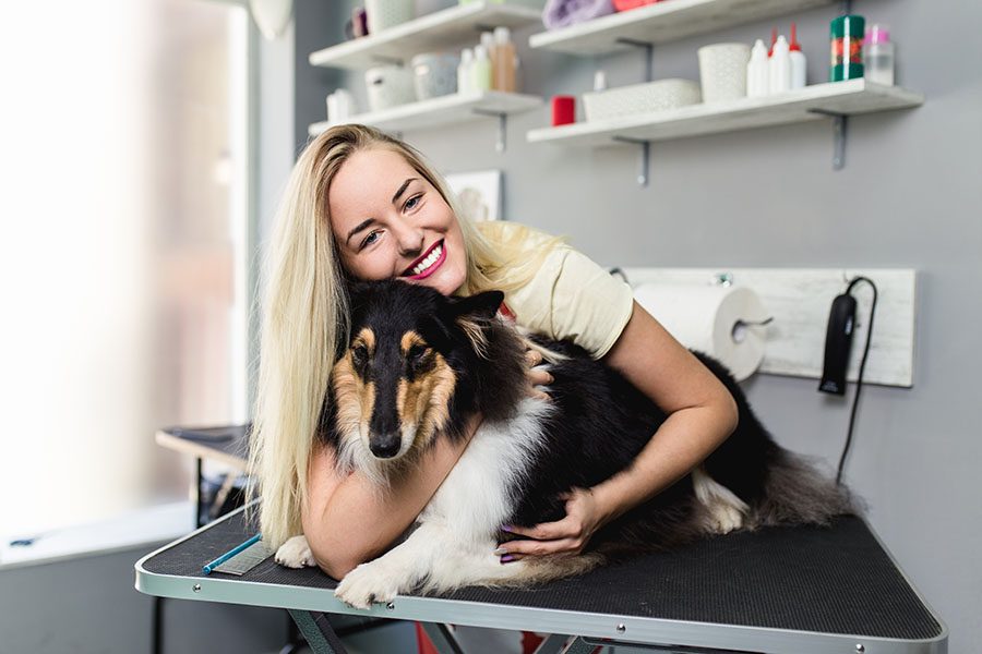 Pet Groomer Insurance - Smiling Dog Groomer Hugging Dog Laying on Grooming Station